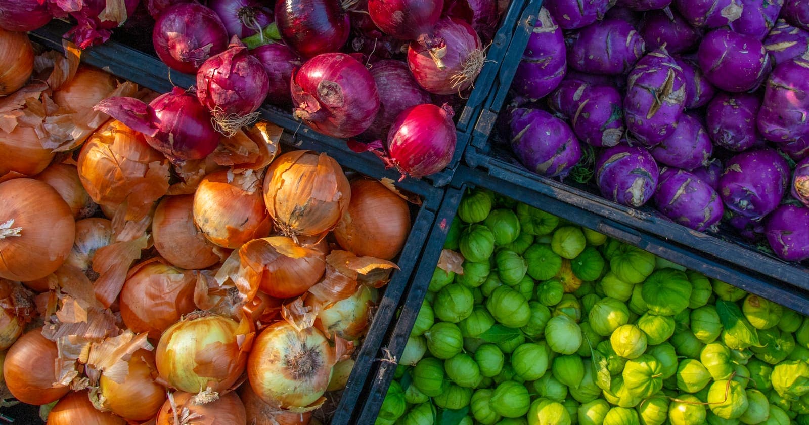 Crates of yellow and red onions with papery skins beside kohlrabi and tomatillos at Corona Farmers Market in Queens, New York