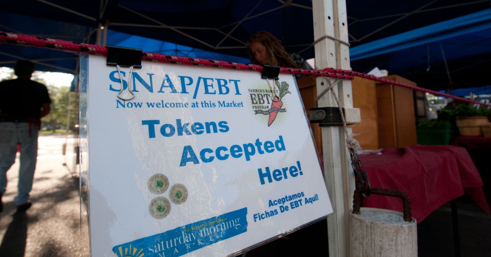 A sign reading SNAP/EBT Now welcome at this Market, Tokens Accepted Here, clipped to a vendor tent at the Saturday Morning Market in St. Petersburg, Florida